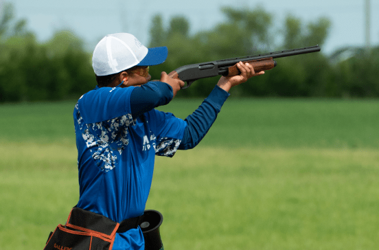 A man in a blue shirt shooting a shotgun in a field.